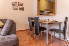 Ein kleines Zimmer mit einer Couch, einem Esstisch mit vier Stühlen, einer Küchenzeile und drei gerahmten Bildern an der Wand.