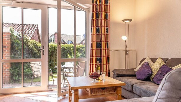 Ein Wohnzimmer mit einer Couch, einem Tisch mit einer Schale und Weingläsern, einem karierten Vorhang und Blick durch große Glasüren auf eine Terrassenanlage.