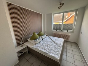 Quarto com cama, mesa de cabeceira, janela e vista para edifícios, e radiador.
