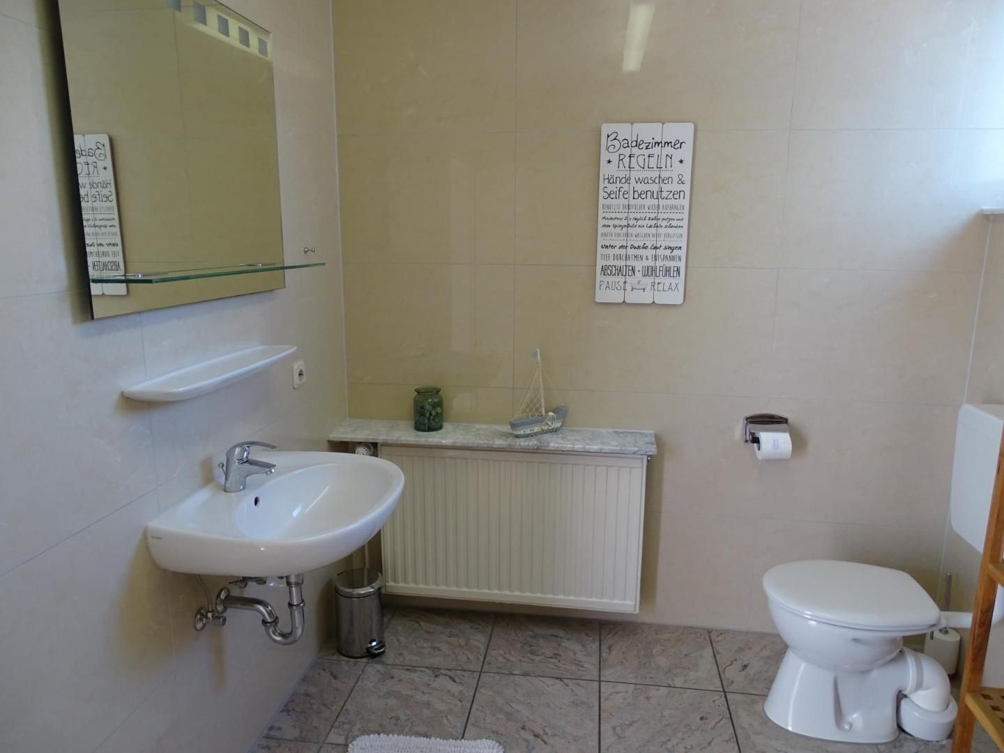 Een badkamer met een witte wastafel, spiegel en toilet. Op de muur hangt een bord met tekst in het Duits.