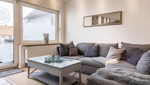 Ein modernes Wohnzimmer mit einem Glastisch, einem grauen Sofa und einem großen Fenster mit Blick auf ein Gebäude.
