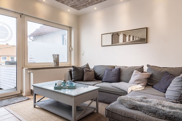 Un salon moderne avec une table basse en verre, un canapé gris et une grande fenêtre donnant sur un immeuble.