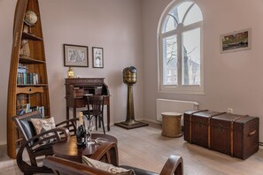 Une pièce comprenant une bibliothèque en bois, un bureau, un globe terrestre, une lampe, une chaise, une table avec une bouteille, une fenêtre et un tableau.
