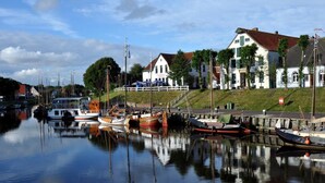 Ein ruhiger Hafen mit mehreren vertäuten Booten, ein weißes Gebäude mit rotem Dach und ein grasbewachsener Uferhang.