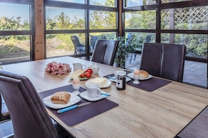 Una mesa de comedor con alimentos para el desayuno, como huevos, pan y una bebida, ubicada en una habitación con ventanas amplias y sillas.