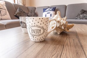 Dos tazas con el texto 'ENJOY', sobre una mesa de madera, junto a una concha decorativa y un sofá al fondo.