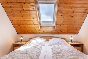 Une chambre avec un plafond en bois incliné, une lucarne, deux tables de chevet avec des lampes et un lit avec une literie blanche.