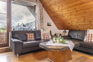 Un salon moderne avec un plafond en bois, des canapés en cuir, une table basse agrémentée d’un arrangement floral et une vue dégagée sur l’extérieur grâce à une grande fenêtre.