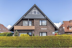 Une maison à deux étages, dotée d’un toit triangulaire imposant, d’un balcon et d’une haie bien entretenue en premier plan.