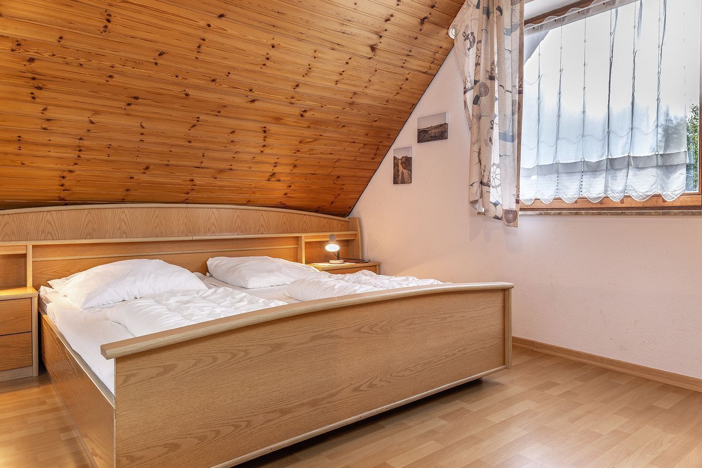 A bedroom with a sloped ceiling, wooden bed frame, bedside tables, and a window with curtains.
