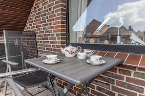Une table dressée avec des tasses à thé blanches et des théières ornées de motifs floraux, une chaise et une fenêtre donnant sur des bâtiments et une sculpture d’oiseau.