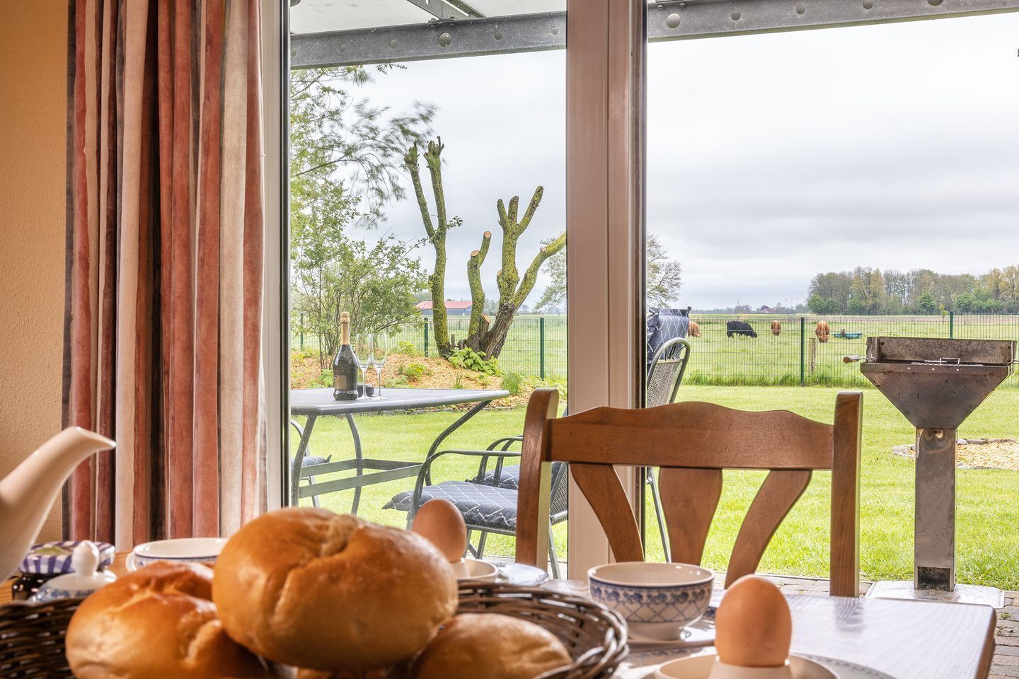 A breakfast table with bread, eggs, and a bottle of wine, overlooking a grassy area with trees and a fence.