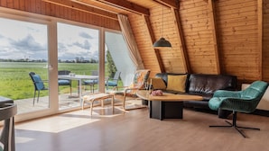 Amplia habitación con un gran ventanal corredizo que da a un espacio exterior, techo de madera y sofá de cuero negro con un sillón color verde azulado.