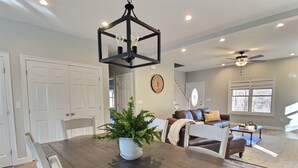A dining area with a wooden table, white chairs, and a potted plant. There is a living room in the background with a sofa, a coffee table, and a window. - Hilltop House (Litchfield)