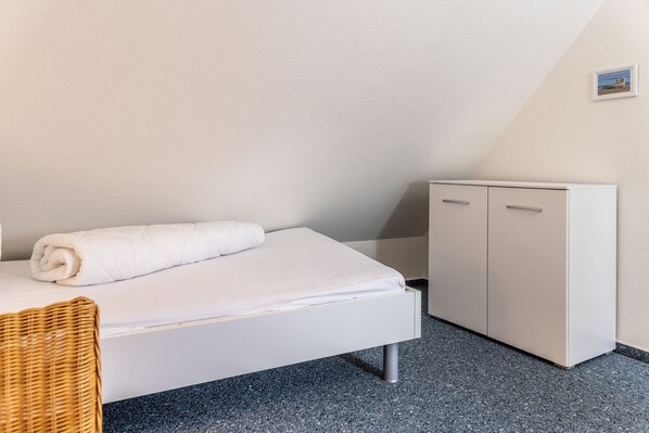 A single bed with a rolled white towel, a white wardrobe, and a wicker chair. - Vacation Home 50009 - Timeless and elegant: This townhouse is truly impressive (Wittmund)