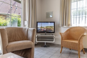 A living room with a beige sofa, a wicker chair, a flat-screen TV on a stand, and a window with curtains. - Vacation Home 50009 - Timeless and elegant: This townhouse is truly impressive (Wittmund)