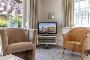 Un soggiorno con un divano color beige, una poltrona in vimini, una televisione a schermo piatto su un mobile e una finestra con tende.