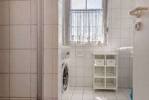 Een kleine wasruimte met een wasmachine, een klein plankje met manden, en een raam met gordijnen.