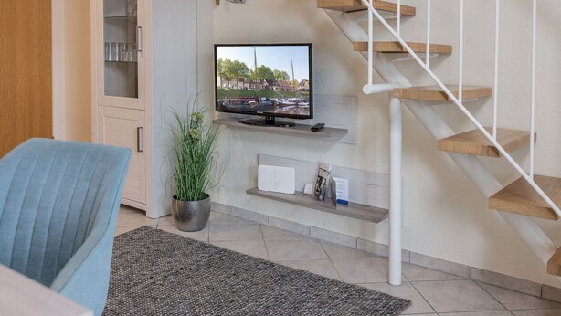 Ein modernes Wohnzimmer mit einem flachbildfernseher an der Wand, einer Holztreppe, einem blauen Sessel, einer Topfpflanze und einem kleinen Regal mit Gegenständen.