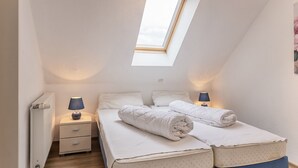 Ein kleines, helles Schlafzimmer mit Dachfenster, einem Bett mit aufgerollten Handtüchern, Nachttischen mit Lampen und einem Heizkörper.