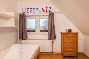 A room with a bed, a wooden dresser, a radiator, a window with curtains, and a sign above the window reading 'LIEGEPLAZ.'