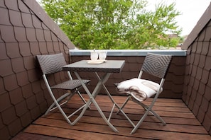 Un petit espace extérieur aménagé avec une table et deux chaises, un coussin sur l’une des chaises, et une vue sur les arbres et les bâtiments en arrière-plan.