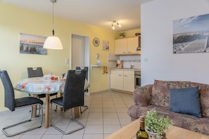 Un coin repas avec une table et des chaises, une cuisine équipée de placards et d’un évier, et un canapé à motifs agrémenté d’une plante posée sur une petite table d’appoint en bois.