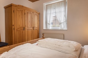 Quarto com guarda-roupa de madeira, cama com roupa de cama branca e janela com cortinas leves.