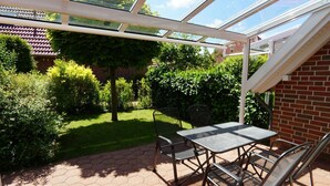 Een overdekt terras met een tafel en stoelen, omgeven door groen en een bakstenen muur.