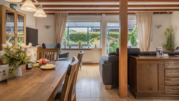 A modern kitchen with wooden countertops, a dining table, and a built-in island. There are pendant lights, a window with a view of greenery, and a cozy seating area with a sofa and armchairs.