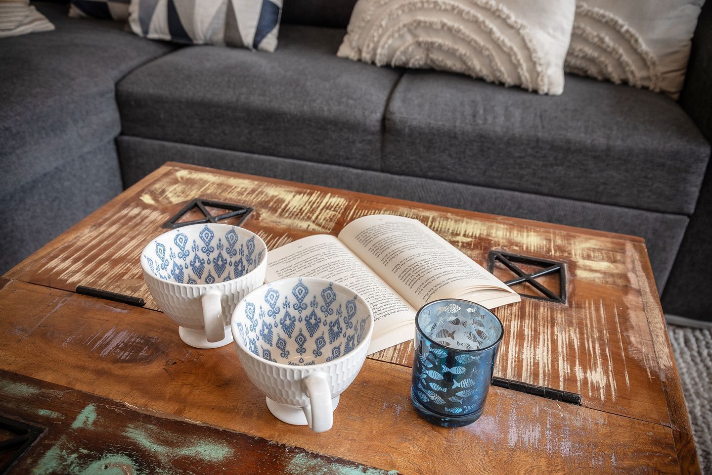 Een houten bijzettafeltje met een boek, twee bekers met een patroon en een glas met vismotieven.