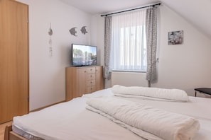 Une chambre à coucher avec un lit, un téléviseur posé sur un meuble en bois, une fenêtre avec des rideaux et un radiateur.