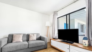 A modern living room with a grey sofa, a flat-screen TV on a wooden console, and a large window with curtains. - Ocean's Sound Hotel (Hollywood)