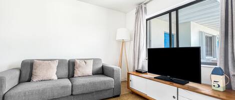 A modern living room with a grey sofa, a flat-screen TV on a wooden console, and a large window with curtains.