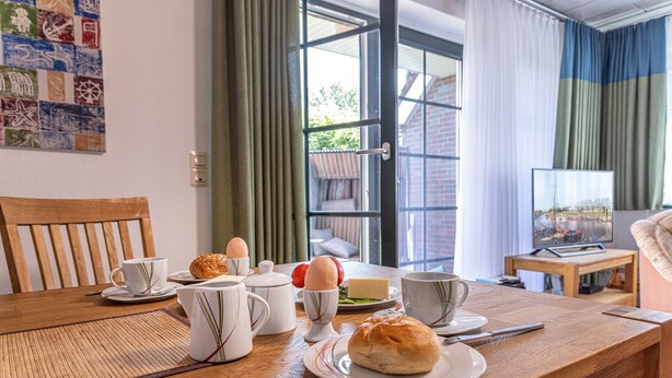 A dining table with breakfast items, a television, and a view of a brick building through a glass door. - Vacation apartment 50036 - Comfortable and cozy: The vacation apartment is located in (Wittmund)