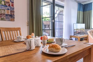 Une table à manger avec des aliments pour le petit-déjeuner, un téléviseur et une vue sur un bâtiment en briques, visible à travers une porte vitrée.