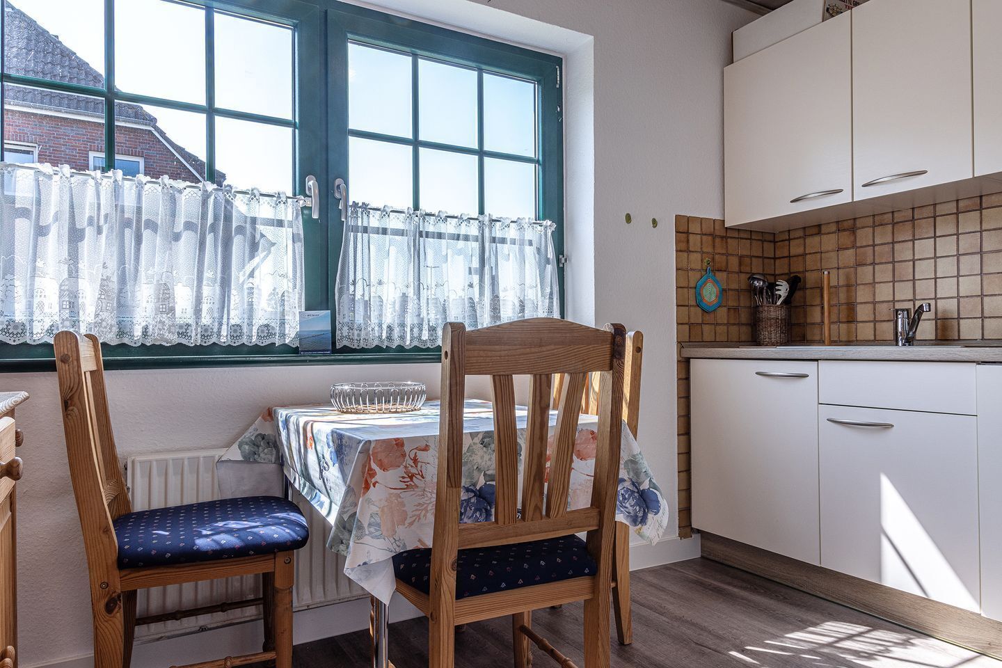 Una cocina con armarios blancos, salpicadero de azulejos y ventana con cortinas de encaje.