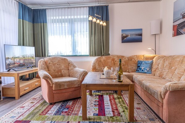 Un salon chaleureux avec un canapé, un fauteuil et une table basse. Un téléviseur est posé sur un meuble en bois, et il y a une fenêtre avec des rideaux.