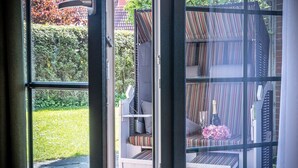 A room with a large window, a bed with striped bedding, a small table with a bottle and flowers, and a view of a garden and a building outside. - Vacation apartment 50036 - Comfortable and cozy: The vacation apartment is located in (Wittmund)
