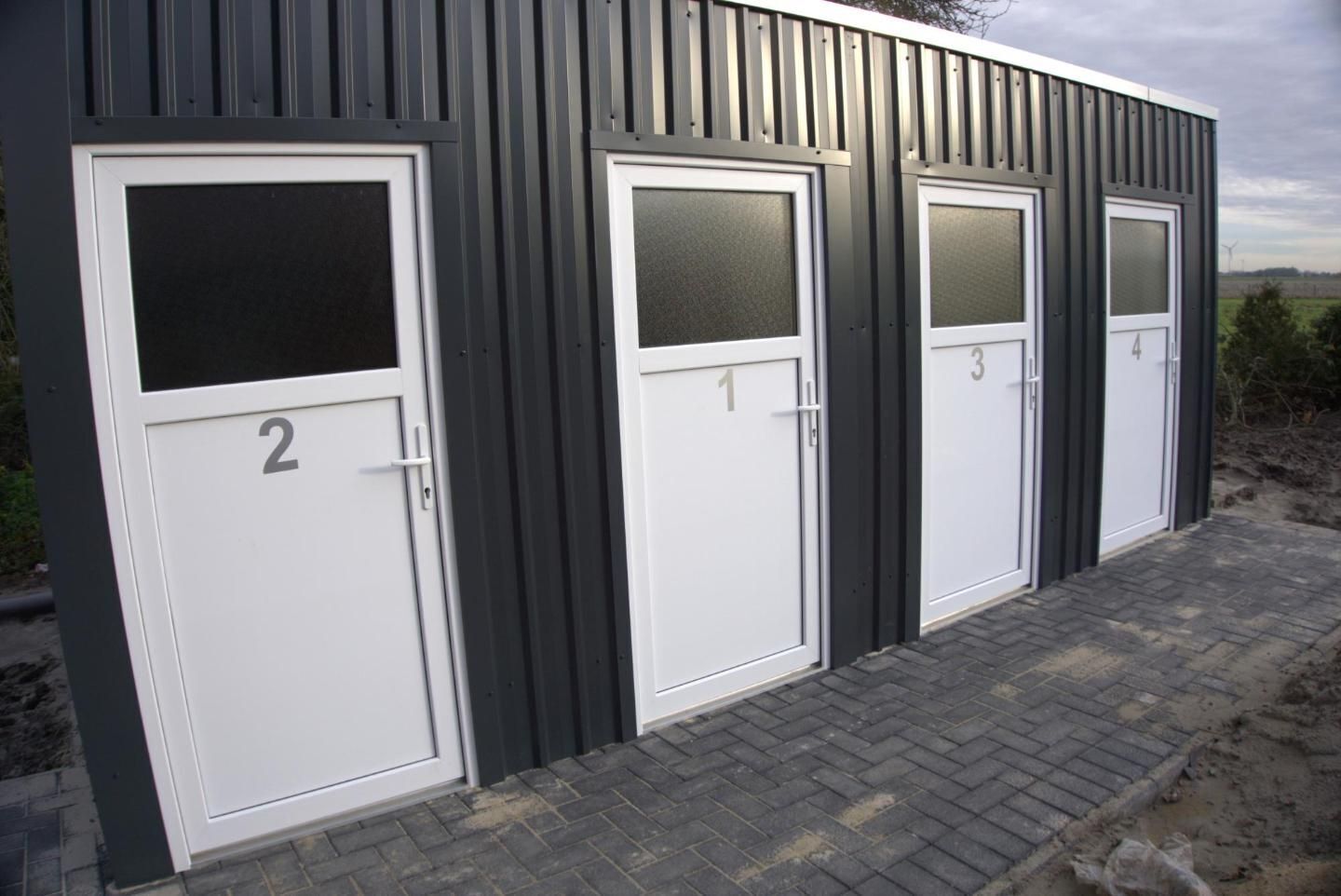 Een rij van vier genummerde deuren, elk met een raam, vast aan een grijs gebouw met een geplaveid pad ervoor.