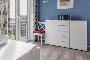 Quarto com cama, guarda-roupa branco, cadeira com almofada vermelha e estante com livros.