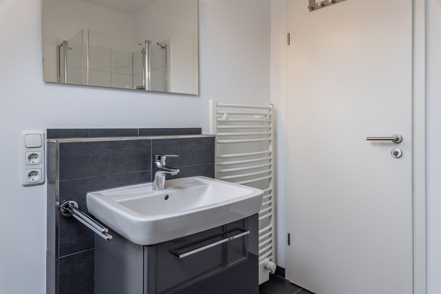 A bathroom with a white sink, dark tiles, a mirror, a towel rack, and a door.