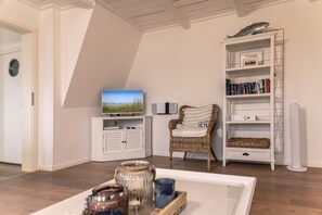 Ett rum med en tv-apparat placerad på ett vitt skåp, en flätstol, en bokhylla och ett litet bord med burkar.