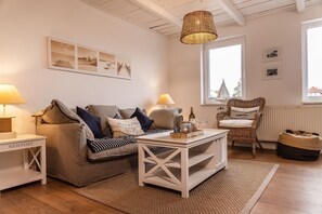Un salon chaleureux avec un canapé, une table basse et un fauteuil en osier. On y trouve deux lampes sur des tables d’appoint, une fenêtre avec vue et un mur orné de tableaux.