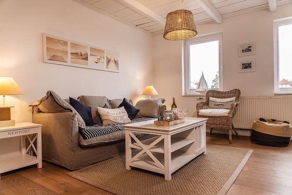 A cozy living room with a sofa, a coffee table, and a wicker chair. There are two lamps on side tables, a window with a view, and a wall with framed pictures. - 50111 Fisherman's Loft - Detached Vacation Home at the Friedrichsschleuse – (Wittmund)