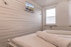 A room with white wooden walls, a bed with white bedding, a window with blinds, and a picture of a harbor scene.