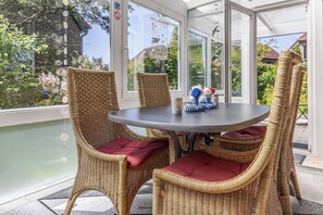 En overdækket terrasse med fletstole og et bord, fuglebur og udsigt til grønne områder og bygninger.