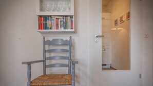 Una silla con asiento y respaldo trenzados, una estantería con libros y gafas, y una puerta que da a un cuarto con espejo y cuadros enmarcados.