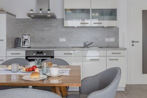 A modern kitchen with white cabinets, a stainless steel range hood, and a built-in oven. There is a dining table with chairs and a breakfast setting.