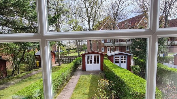 A view through a window of a well-maintained garden with a pathway leading to a small red building, surrounded by neatly trimmed hedges and trees. - Strandkrabbe - Holiday home Strandkrabbe - close to the beach, 3 bedrooms, garden (Wyk auf Föhr)